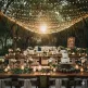 Decoração de casamento rústico e simples com folhagens verdes, luzes de varal e móveis de madeira, criando um ambiente aconchegante.